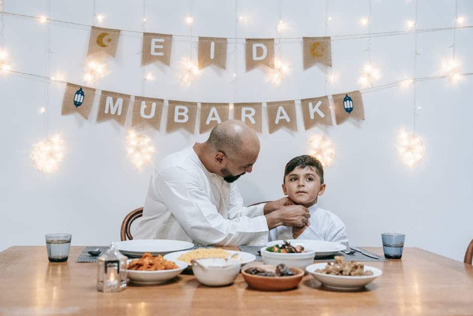 A father adjusts his son's shirt as they sit together celebrating Eid indoors with festive lights and food.